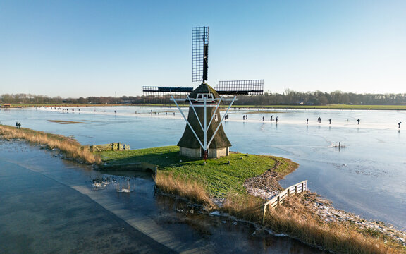 Aerial view of Ypey Mole windmill surrounded by a frozen landscape with people ice skating under a clear blue sky in Ryptsjerk, Friesland, Netherlands.