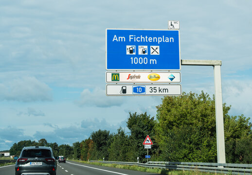 Germany - Aug 22, 2026: Cantilever sign for the Am Fichtenplan service area at 1000 meters beside the motorway with a dark car passing below and green trees lining the grass verge