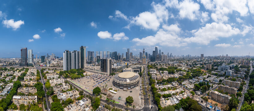 Aerial view of Menora Mivtachim Arena surrounded by urban buildings, parking lots, and city streets under a blue sky with scattered clouds in Tel Aviv-Yafo, Tel Aviv District, Israel.
