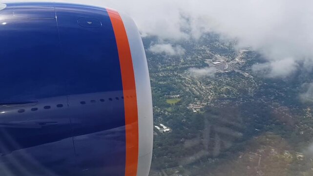 Airplane jet engine viewed from porthole during flight descent. Aircraft flying over city and green landscape through white clouds.