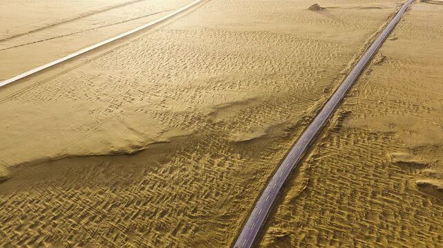 Aerial view of a paved highway crossing an arid desert with grid sand barriers and rippled dunes under bright sunlight.