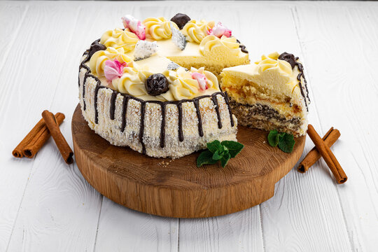 Sliced layered cake with cream rosettes on wooden board. Round sweet pastry decorated with dark chocolate drips and prune toppings next to cinnamon sticks.