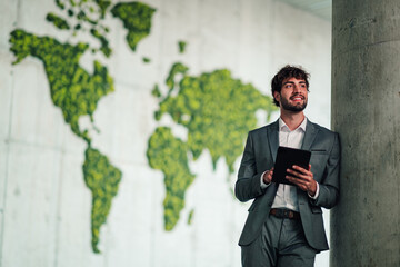 Obraz na płótnie Canvas Businessman holding a tablet, smiling and looking up, standing in front of a green moss world map wall
