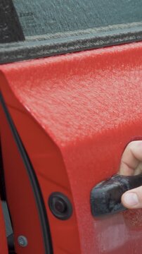 Male driver hand pulls ice covered door handle and opens door of red car despite thick frozen coating. Solid glaze ice covers vehicle from roof to door. Subzero temperature and severe winter weather
