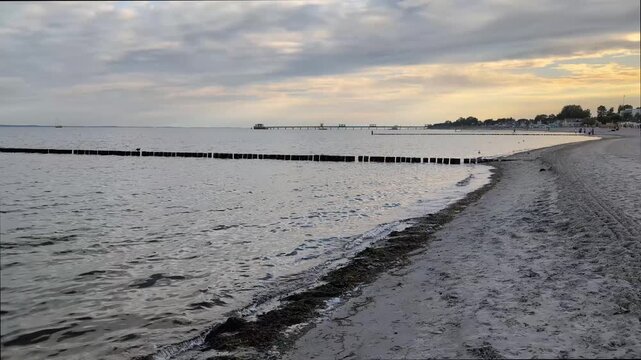 Calm Baltic Sea coast at sunset with wooden breakwater (Groyne), sandy shore, and pier under a cloudy sky