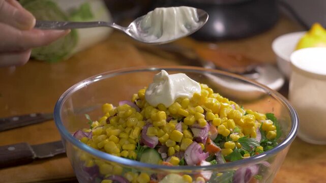 Hand pours mayonnaise over boiled corn, red onion, imitation crab meat, fresh cucumber and minced parsley. Homemade salad combines natural ingredients, savory taste and varied texture