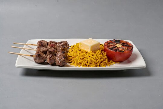 Chelo Kebab on white rectangular plate features grilled meat skewers, yellow rice, small block of butter, grilled tomato. Concept of grilled meat with saffron rice butter.