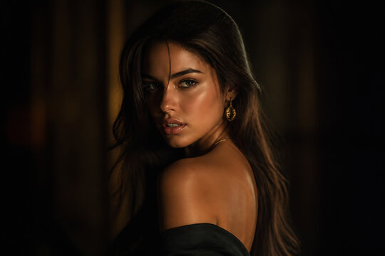 A beautiful woman with long dark hair is posing in a dimly lit, moody setting.