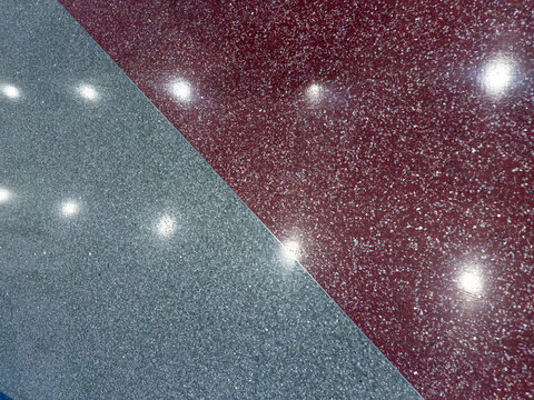 Close-up of glossy terrazzo flooring where a deep red section meets a blue-gray section along a diagonal seam, with ceiling lights reflecting off the polished surface