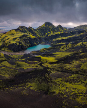 Aerial view of Icelandic Highlands with vibrant green moss-covered volcanic terrain, dark basalt rock formations, and a turquoise glacial lake under a dramatic cloudy sky Iceland.