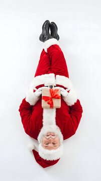 Santa claus upside down holding a wrapped christmas gift box with red ribbon wrapped present white background celebration surprise