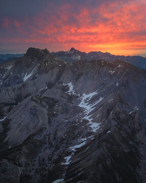 Aerial view of the French Alps at sunset with dramatic orange and red clouds over rugged snow-capped mountain peaks and rocky slopes in France.