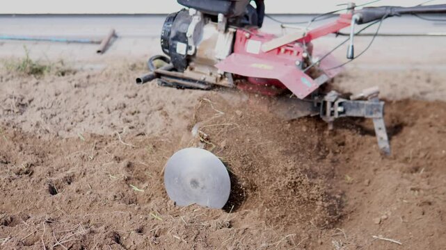 Red and black farm cultivator disturbs dry soil beside paved edge, throwing dust and loose earth across rough foreground, with farm, agriculture, tilling, soil detail, showing dust, cultivator