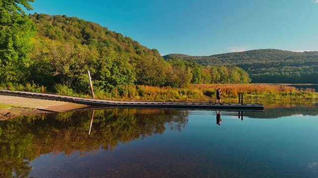 Traveler walking on a dock at Sherman Reservoir Boat Launch in Vermont, USA, surrounded by calm water, autumn hills, reflections, and scenic outdoor landscape. Scenic drone view. 