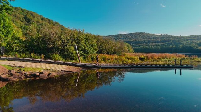 Traveler walking on a dock at Sherman Reservoir Boat Launch in Vermont, USA, surrounded by calm water, autumn hills, reflections, and scenic outdoor landscape. Scenic drone view. 4K.