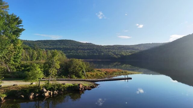 Peaceful nature and boating recreation. Scenic calm water left to right drone view at Sherman Reservoir Boat Launch, Vermont, USA, with forested mountains, tree reflections and clear sky. 4K.