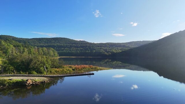 Peaceful nature and boating. Scenic water static drone view at Sherman Reservoir Boat Launch, Vermont, USA, with forested mountains, tree reflections and clear sky. Traveler walking on a dock. 4K.