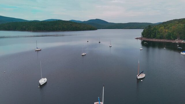 Aerial view of sailboats moored on calm Harriman Reservoir, Vermont, USA, framed by forested hills and distant mountains beneath a soft, overcast sky. Slow dolly shot.