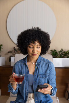 African American woman sitting at home on chair near mirror holding teacup checking smartphone