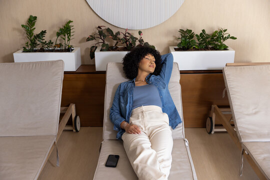 African American woman reclining on center padded lounger at spa lounge, smartphone resting