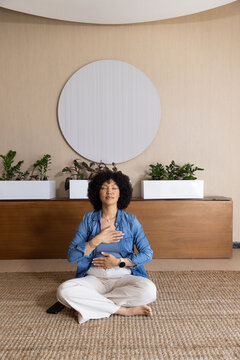 African American woman sitting with legs crossed on jute rug in calm room, breathing, phone nearby