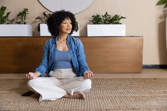 African American woman sitting on woven rug at home, wearing denim shirt, smartphone nearby