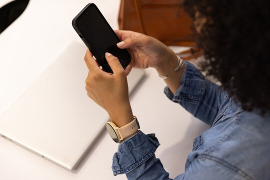 African American woman wearing denim jacket using smartphone at white desk with laptop, copy space