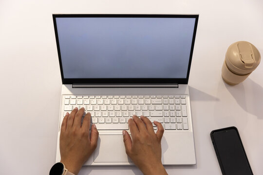 Woman typing on white laptop at tidy desk with smartwatch, black smartphone and beige travel cup