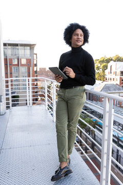 African man standing on rooftop walkway leaning on white rail in black turtleneck using tablet