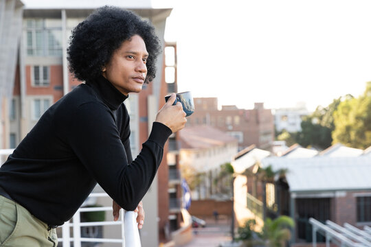 African American man in black turtleneck leaning on balcony rail with dark mug, gazing at rooftops