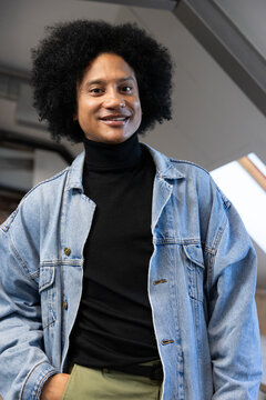 Man standing in loft facing camera under skylight, wearing denim jacket and black turtleneck