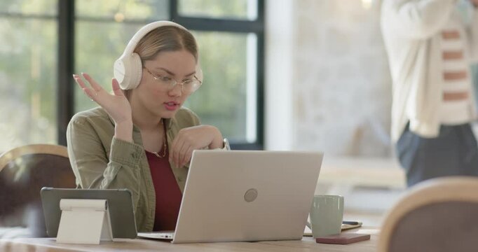 Asian adult woman adjusting headphones, responding to incoming call, gesturing at laptop in office