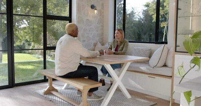 Diverse couple conversing at dining nook, man gesturing and sipping green mug near smartphone