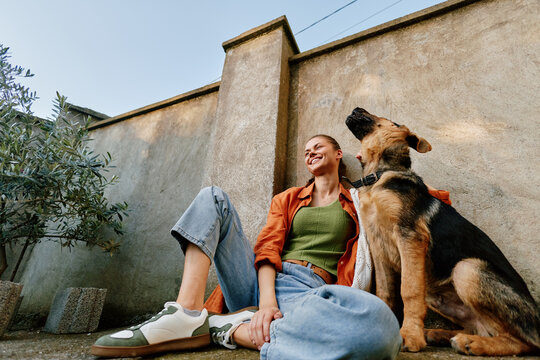 Smiling young woman sits on the ground by a concrete wall with her german shepherd dog, enjoying a candid moment of friendship and trust outdoors.