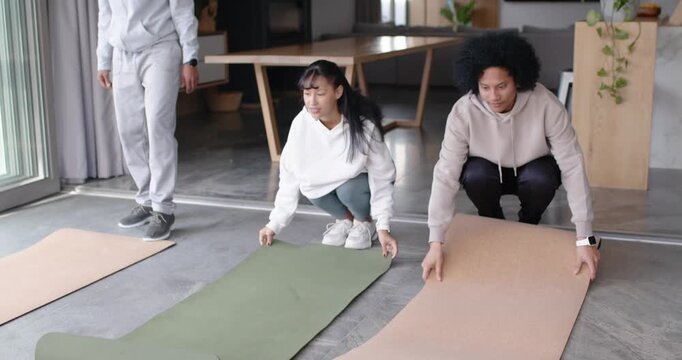 Diverse friends unrolling three mats in living room, standing, warming up in activewear for workout