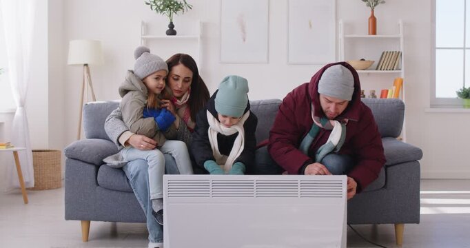 Portrait of frozen young family with kids sitting on sofa in the living room in winter hats and coats at home and trying to warm on electric heater. Heating problems concept. 4k video.