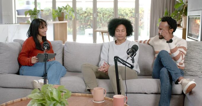Diverse cohosts and guest talking on couch as right host speaking gesturing using tablet and mics