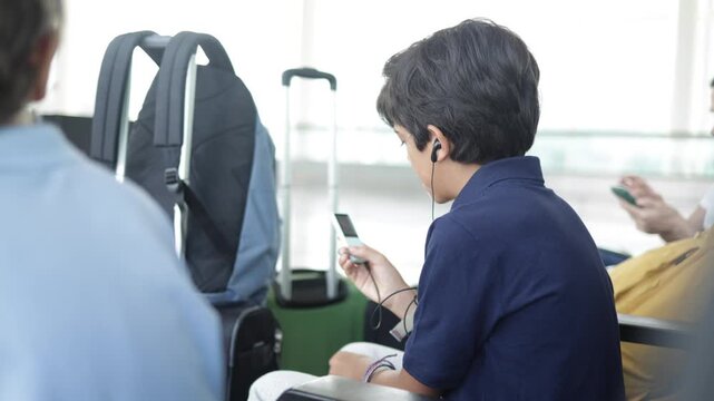 Young boy with earphones listening to music on his mp3 player while waiting at the airport terminal gate before boarding, his luggage and backpack ready for the trip