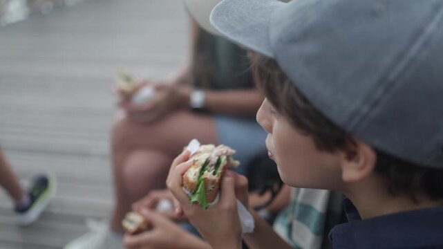 Young boy with a baseball cap eating a sandwich with his family outdoors on a sunny day. A family enjoying a meal together on a new york boardwalk, representing urban lifestyle