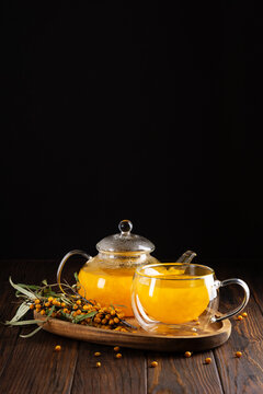 Glass teapot and cup filled with bright orange sea buckthorn berry juice served on a wooden tray with fresh berries and leaves against a dark background