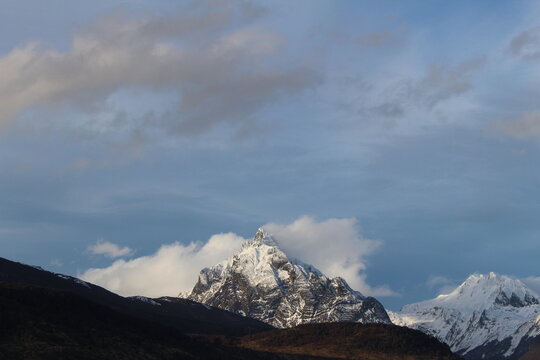Wide landscape photo of the snow-capped peak of Mount Olivia partially veiled by passing clouds with warm light illuminating the snow near Ushuaia, Argentina.