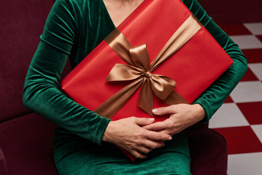 Woman in elegant green velvet dress holding large red christmas gift with gold ribbon.