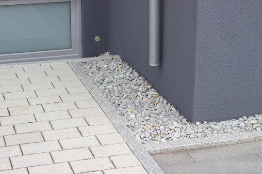 Modern building corner featuring gray stone gravel landscaping alongside a paved walkway and a vertical drainage pipe against a textured wall