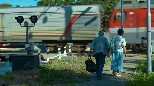 An elderly couple is walking toward the railway station. Red warning lights are flashing to signal the approach of a train. The train passes by the people.