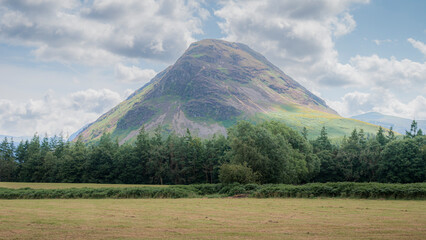 Fototapeta premium A large, grassy mountain looms under a cloudy sky, with a dense forest in the foreground.