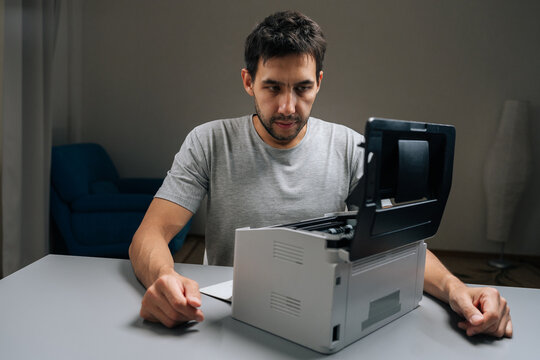 Serious puzzled technician with concentrated expression inspecting laser printer with cover open, attempting to repair it sitting at desk in dark repair shop.