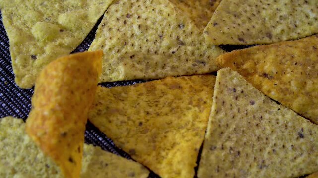 Scattered triangular corn nacho chips with spices rotating on dark textured mat, top view