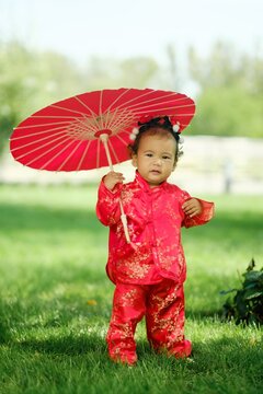 Asian baby girl in traditional red suit holding umbrella standing on green grass outdoors