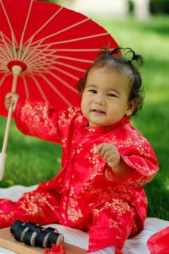 Asian baby in traditional red costume with umbrella, sushi rolls and lantern on picnic blanket