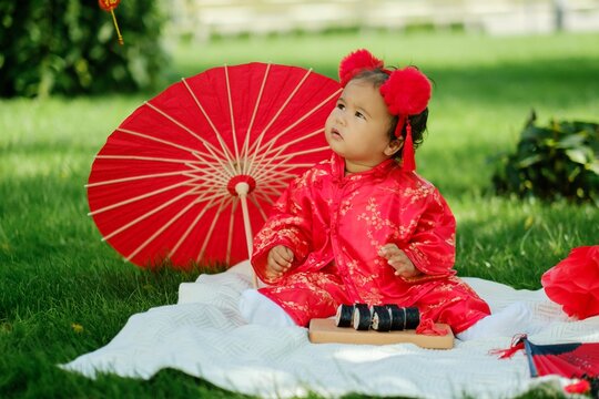 Asian baby in traditional red costume with umbrella, sushi rolls and lantern on picnic blanket